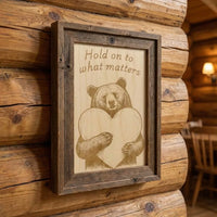 Wooden framed picture with bear holding a heart and text 'Hold on to what matters' on a wooden wall.