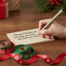 Hand writing a Christmas message on a card with ribbons and a pen on a wooden surface.