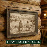 Framed artwork of a couple sitting on a bench by a lake, with text 'You Stayed', on a wooden wall.